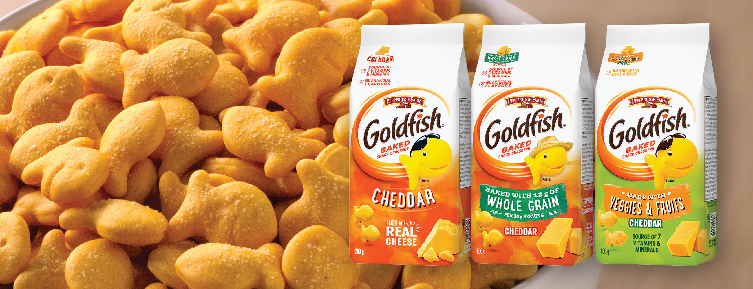 Goldfish® Cheddar, Whole Grain and Veggies & Fruits packaging with bowl of Goldfish® crackers in background